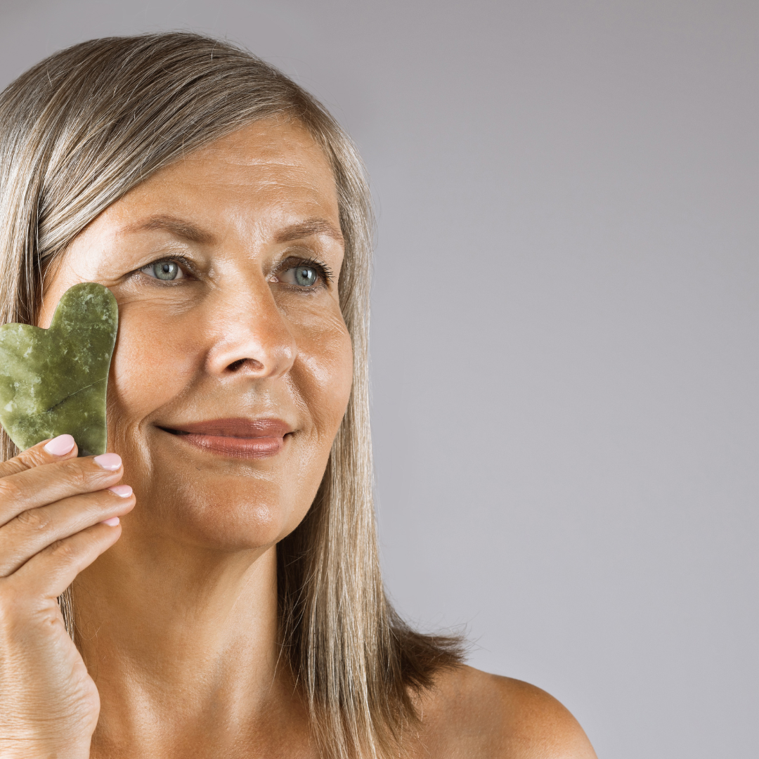 A woman with grey hair holding green gua sha tool next to her face