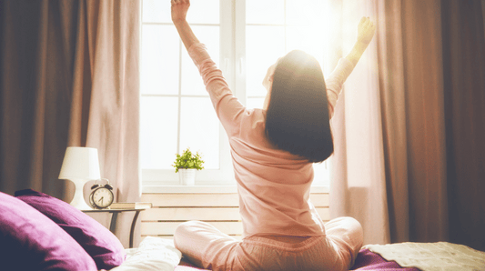 A woman stretching her arms while sitting on bed. Morning sun shining through the window.